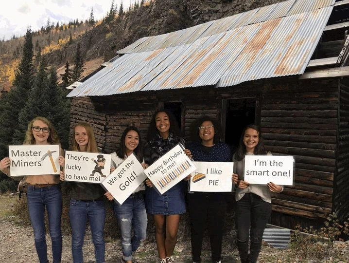 Group of smiling kids holding playful signs in front of a rustic cabin, celebrating a birthday party with a fun, themed escape room adventure at Think in a Box in San Angelo.